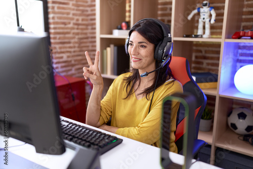 Middle age hispanic woman playing video games using headphones smiling with happy face winking at the camera doing victory sign. number two.