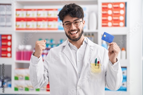 Hispanic man with beard working at pharmacy drugstore holding credit card screaming proud, celebrating victory and success very excited with raised arms