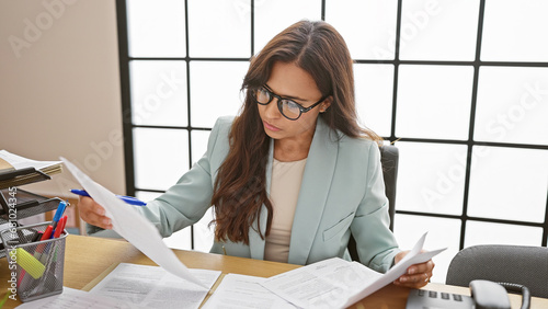 Tablou pe pânză Young, beautiful hispanic woman worker in business attire, concentrated and focused on reading documents while sitting at the office table, embodying elegance and professionalism indoors