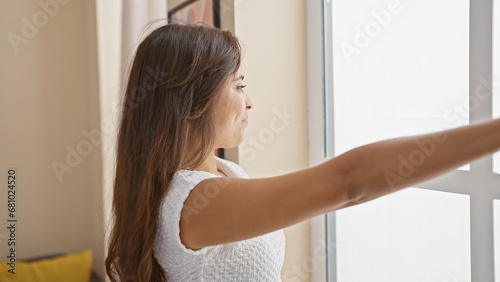 Joyful young hispanic woman, laughing and throwing glimpses outside, stands by the living room window at home with confident smile