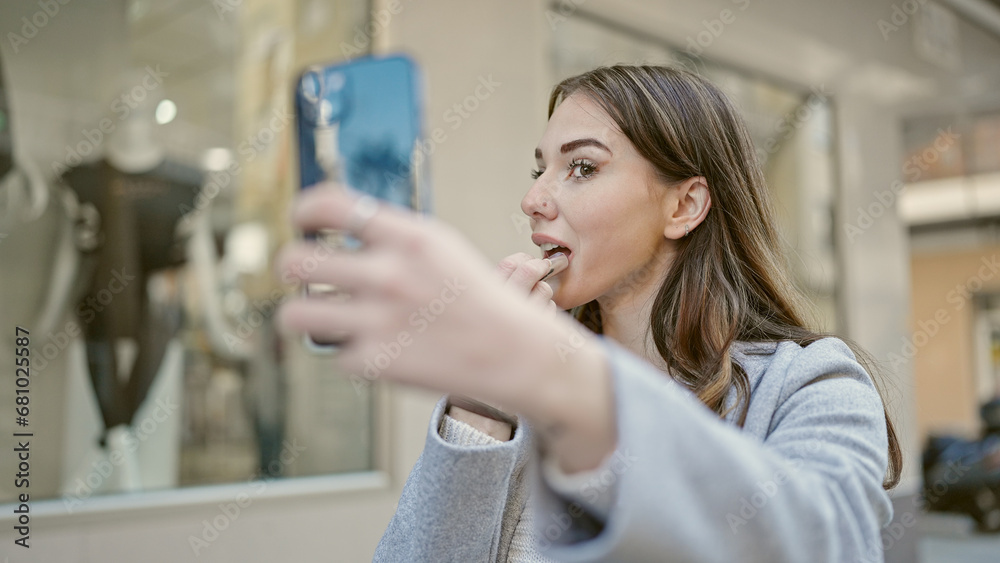 Young hispanic woman using smartphone as a mirror applying lipstick at street