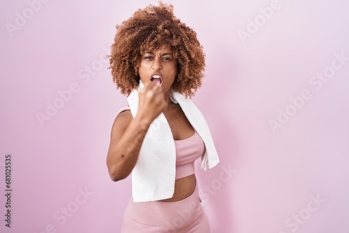 Young hispanic woman with curly hair wearing sportswear and towel angry and mad raising fist frustrated and furious while shouting with anger. rage and aggressive concept.