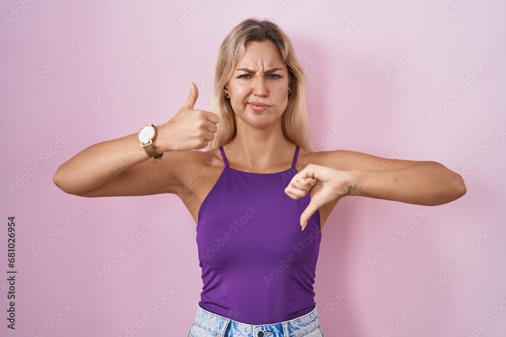 Young blonde woman standing over pink background doing thumbs up and down, disagreement and agreement expression. crazy conflict
