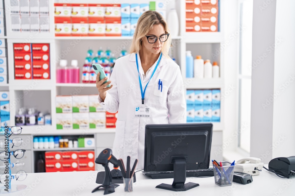 Young blonde woman pharmacist using smartphone and computer at pharmacy