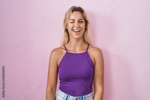 Young blonde woman standing over pink background winking looking at the camera with sexy expression, cheerful and happy face.