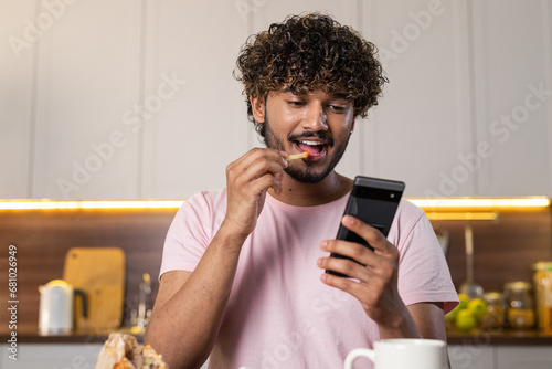 A mestizo guy in the kitchen in subdued lighting, holds a phone and potato fries in hands, eats seductively with pleasure and reads romantic message in the screen. A young american or african man photo