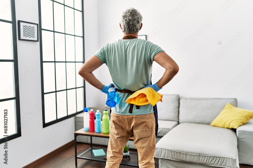 Middle age grey-haired man holding cloth and sprayer standing on back view at home