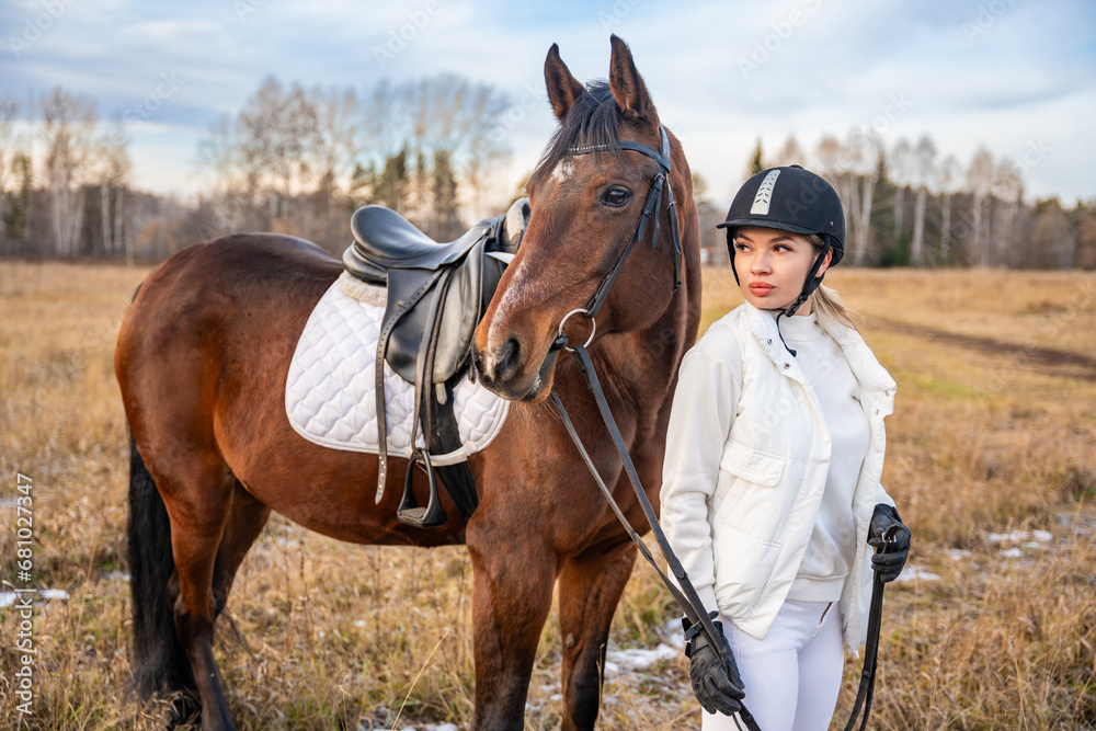 Blond professional female jockey standing near brown horse in field. Friendship with horse Blond professional female jockey standing near brown horse in field. Friendship with horse