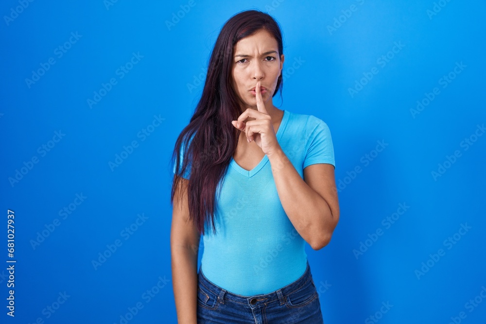 Young hispanic woman standing over blue background asking to be quiet with finger on lips. silence and secret concept. Young hispanic woman standing over blue background asking to be quiet with finger on lips. silence and secret concept.