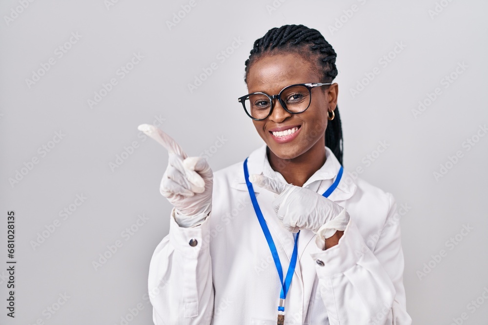 Φωτογραφία African woman with braids wearing scientist uniform smiling and looking at the camera pointing with two hands and fingers to the side