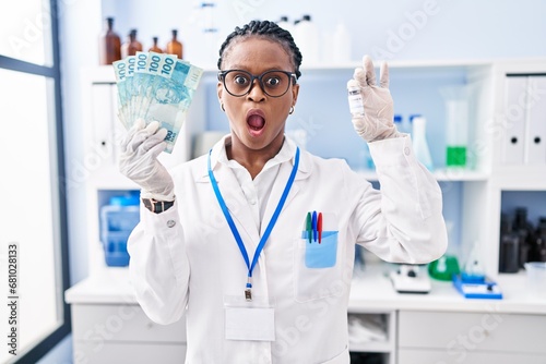 African woman with braids working at scientist laboratory holding money afraid and shocked with surprise and amazed expression  fear and excited face.