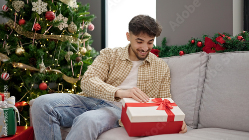 Young arab man unpacking gift with surprised face sitting by christmas tree at home