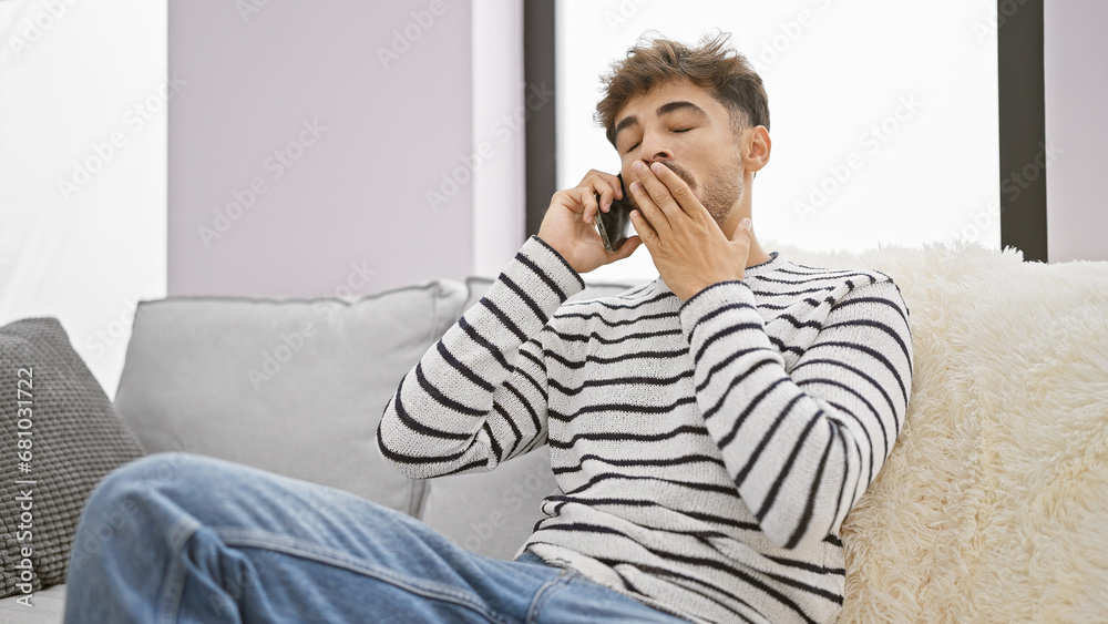 Exhausted young arab man is sitting at home on the sofa, absorbed in a tiresome call, yawning while talking on his smartphone indoors