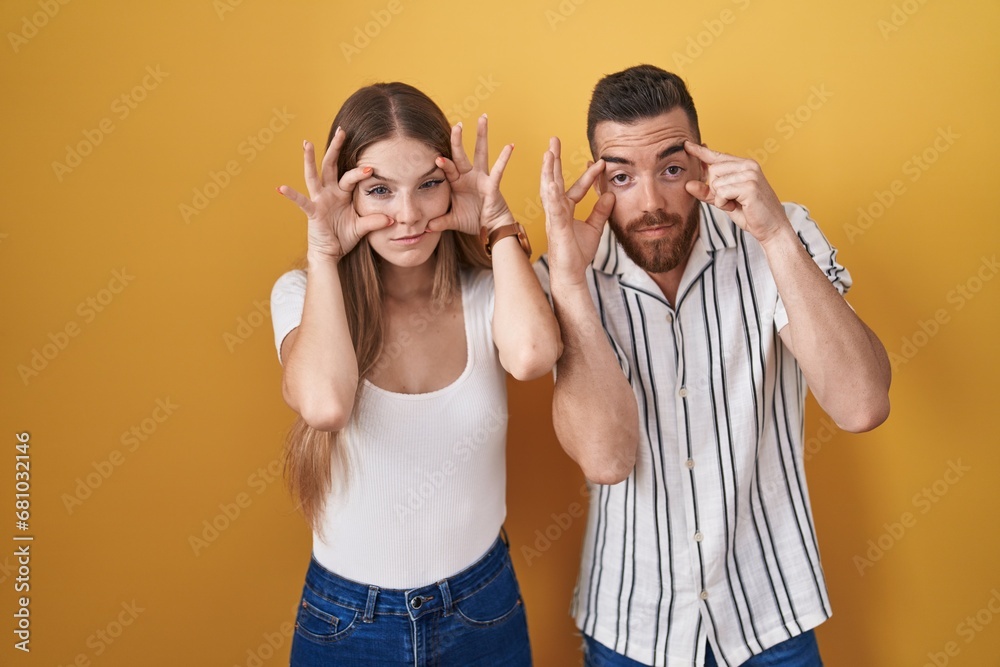 Young couple standing over yellow background trying to open eyes with fingers, sleepy and tired for morning fatigue Young couple standing over yellow background trying to open eyes with fingers, sleepy and tired for morning fatigue