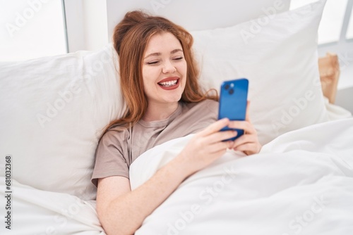 Young redhead woman using smartphone lying on bed at bedroom