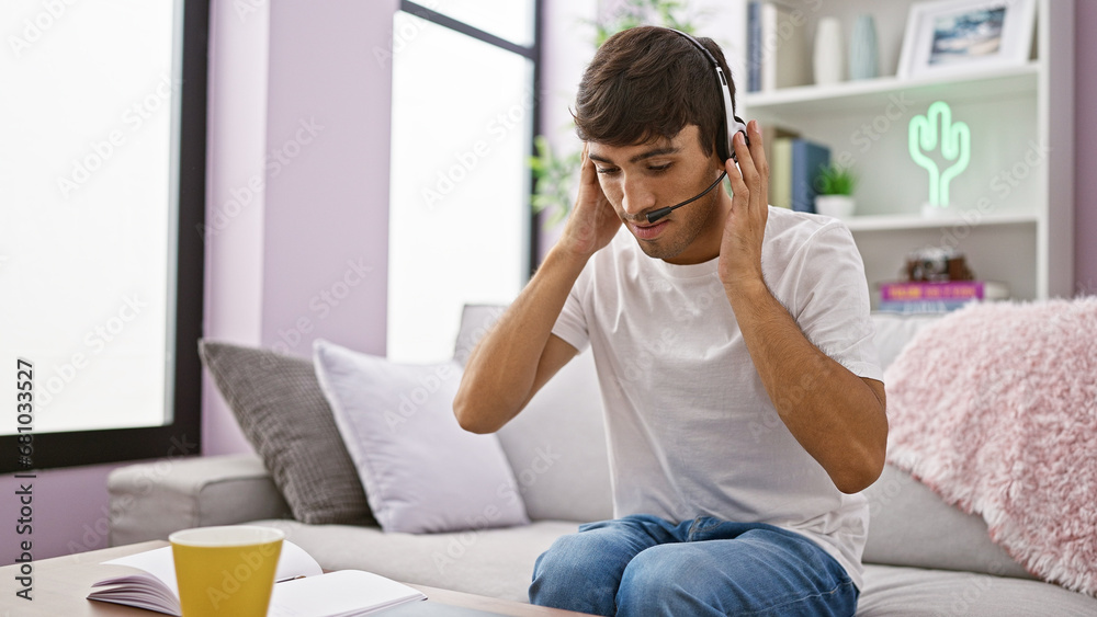 Dedicated young hispanic man, a handy call center agent working from home, settled on his comfy sofa. wearing headphones while sipping coffee, immersed in technology and online support. Dedicated young hispanic man, a handy call center agent working from home, settled on his comfy sofa. wearing headphones while sipping coffee, immersed in technology and online support.