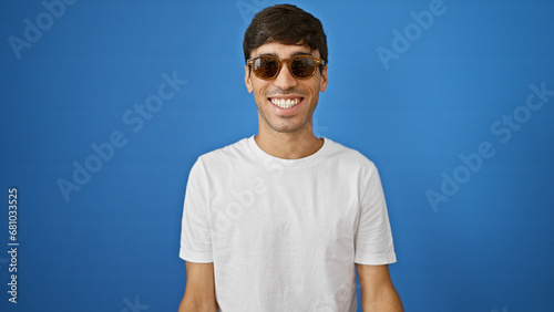 Cheerful young hispanic man wearing sunglasses, exuding confidence and positivity, standing against an isolated blue background while enjoying a cool, casual lifestyle. photo