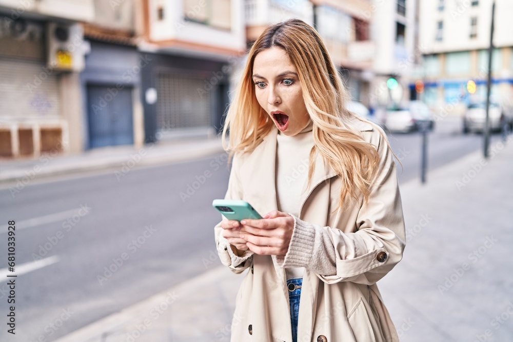 Young blonde woman using smartphone with surprise expression at street