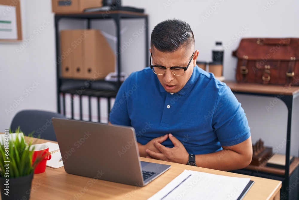 Young hispanic man working at the office with laptop with hand on stomach because nausea, painful disease feeling unwell. ache concept. Young hispanic man working at the office with laptop with hand on stomach because nausea, painful disease feeling unwell. ache concept.