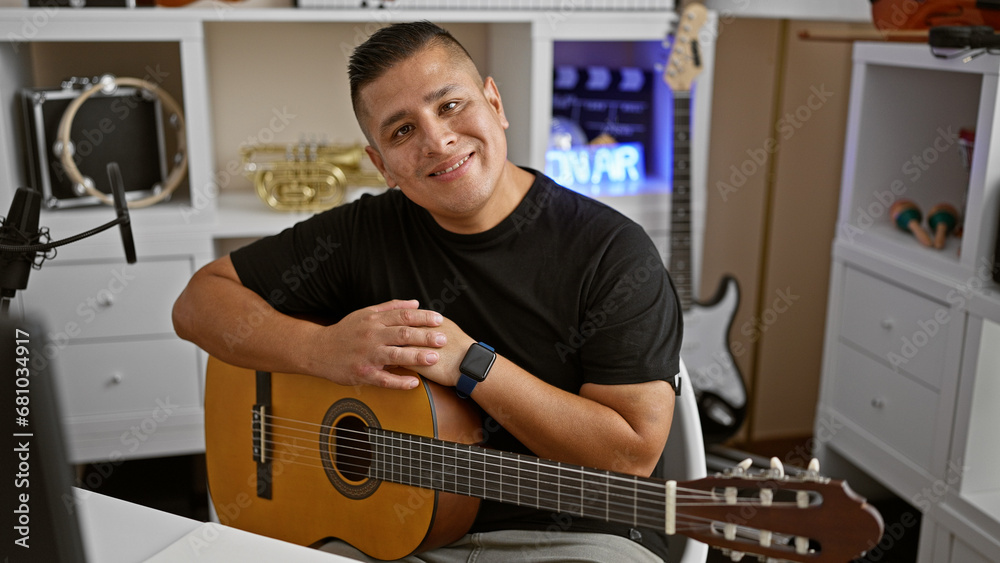Obraz premium Handsome young latin man, a confident musician, holding his classic guitar and smiling radiantly in a music studio, evoking the melody of his acoustic performance