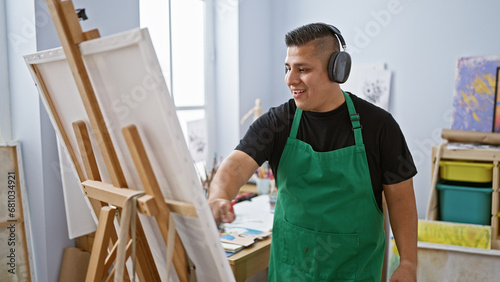 Smiling, handsome latin guy with headphones, utterly absorbed in his music while beautifully bringing his drawing to life at an inspiring university art studio.
