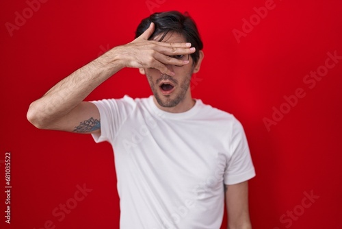 Young hispanic man standing over red background peeking in shock covering face and eyes with hand, looking through fingers with embarrassed expression.