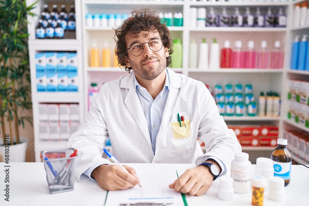 Hispanic young man working at pharmacy drugstore smiling looking to the side and staring away thinking.