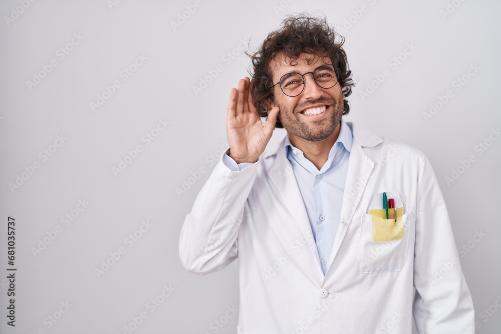 Hispanic young man wearing doctor uniform smiling with hand over ear listening an hearing to rumor or gossip. deafness concept. Hispanic young man wearing doctor uniform smiling with hand over ear listening an hearing to rumor or gossip. deafness concept.