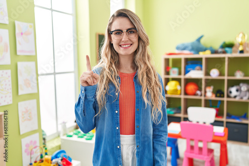 Young caucasian woman working as teacher at kindergarten pointing finger up with successful idea. exited and happy. number one.