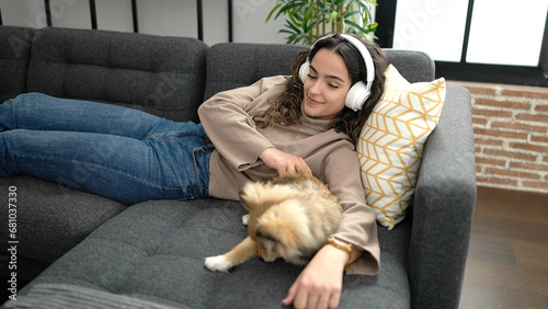Young hispanic woman with dog listening to music lying on sofa at home