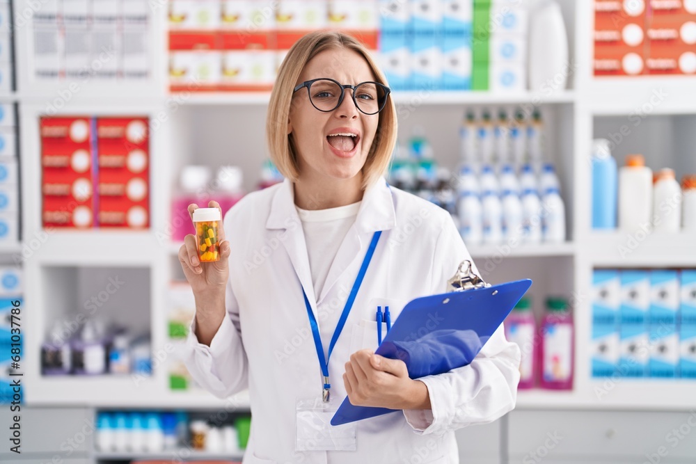 Young caucasian woman working at pharmacy drugstore holding pills angry and mad screaming frustrated and furious, shouting with anger. rage and aggressive concept.