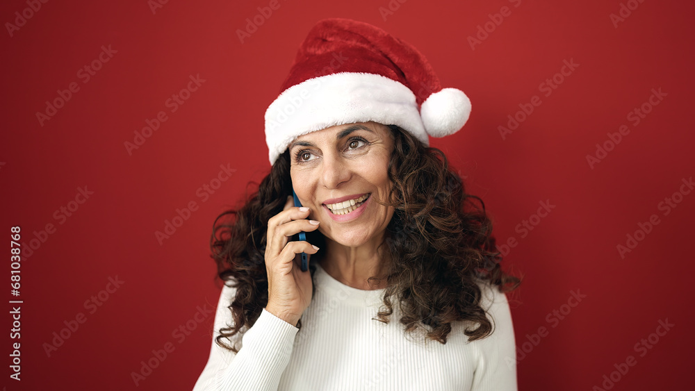 Middle age hispanic woman wearing christmas hat talking on smartphone over isolated red background