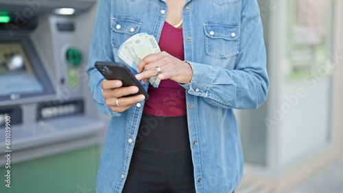 Young beautiful hispanic woman holding chilean pesos banknotes using smartphone at bank teller