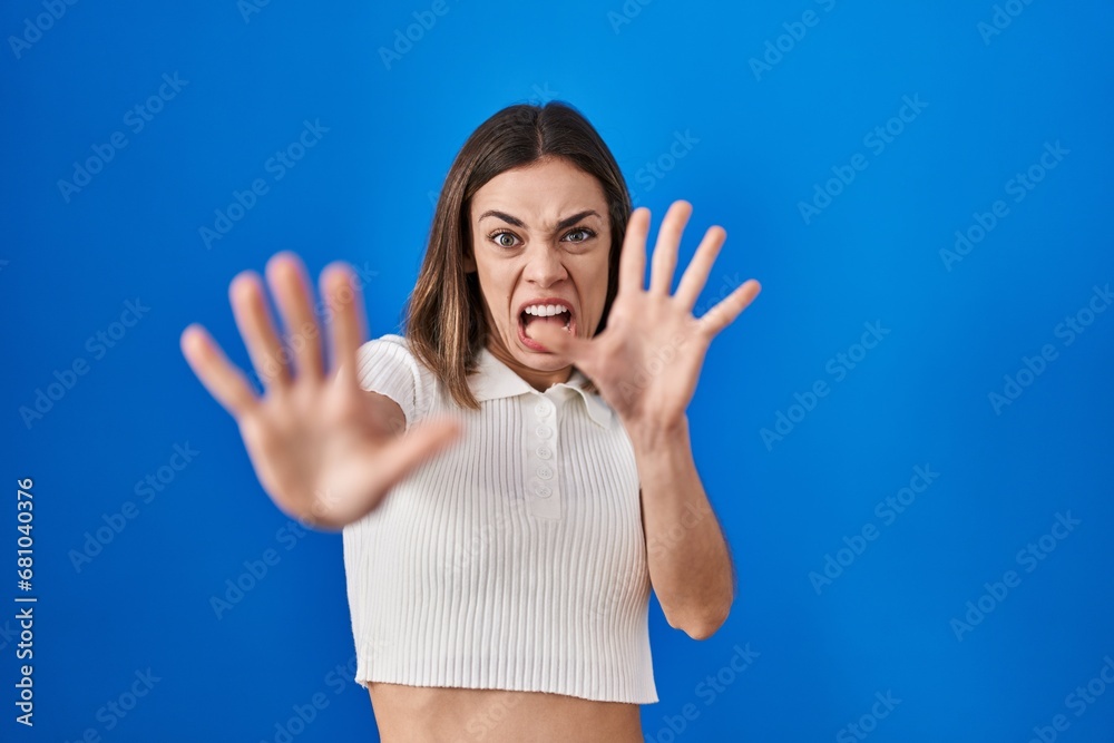 Hispanic woman standing over blue background afraid and terrified with fear expression stop gesture with hands, shouting in shock. panic concept. Hispanic woman standing over blue background afraid and terrified with fear expression stop gesture with hands, shouting in shock. panic concept.