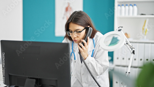 Young beautiful hispanic woman doctor using computer talking on telephone at clinic