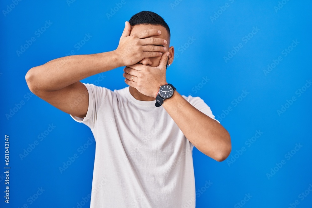 Young hispanic man standing over blue background covering eyes and mouth with hands, surprised and shocked. hiding emotion