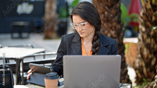 Young beautiful hispanic woman business worker using laptop and touchpad sitting on table at coffee shop terrace