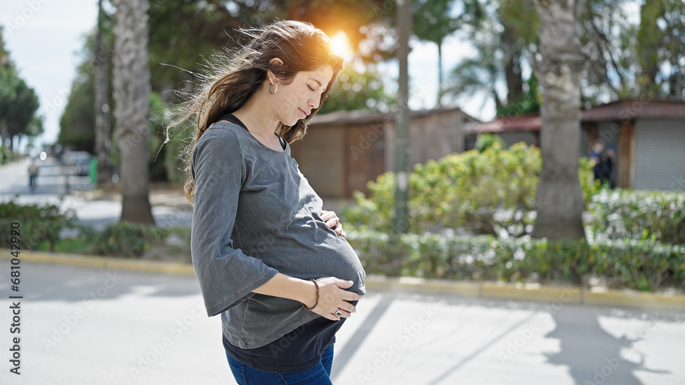 Young pregnant woman standing with serious expression touching belly at park