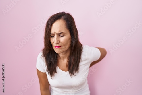 Middle age brunette woman standing over pink background suffering of backache, touching back with hand, muscular pain