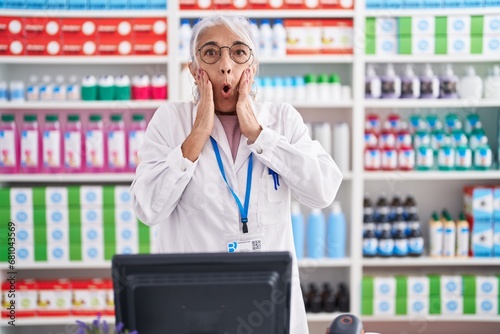 Middle age woman with tattoos working at pharmacy drugstore afraid and shocked, surprise and amazed expression with hands on face