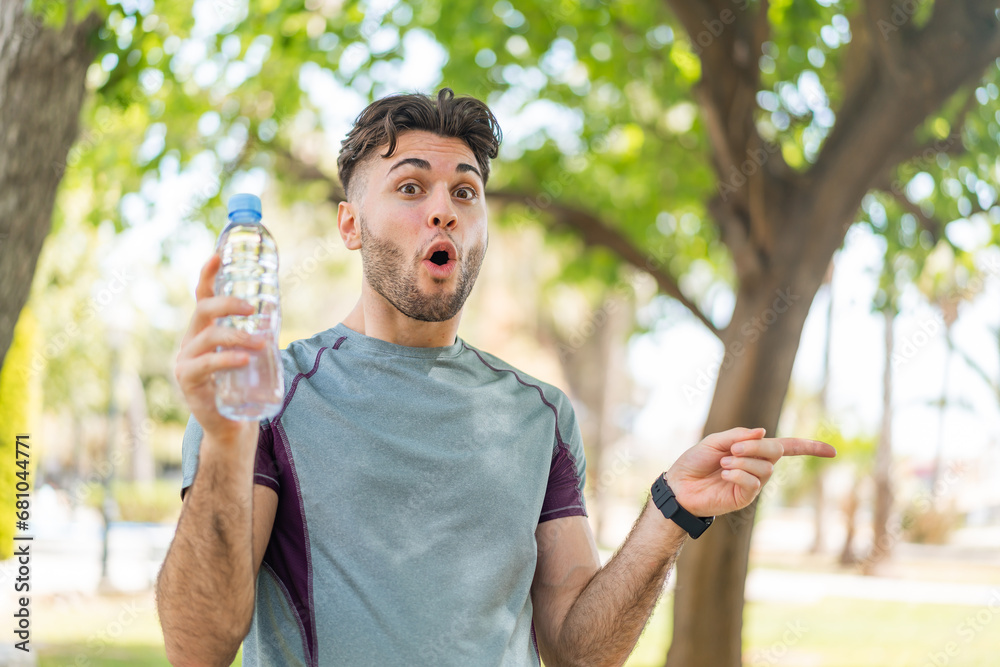 Obraz premium Young handsome sport man with a bottle of water at outdoors surprised and pointing side