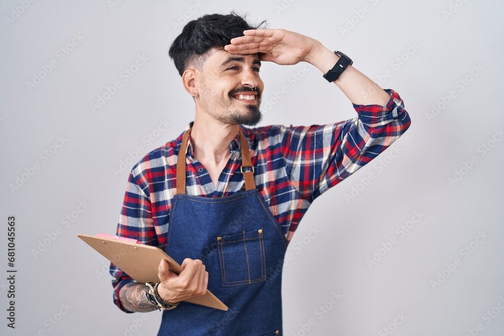 Young hispanic man with beard wearing waiter apron holding clipboard very happy and smiling looking far away with hand over head. searching concept. Young hispanic man with beard wearing waiter apron holding clipboard very happy and smiling looking far away with hand over head. searching concept.