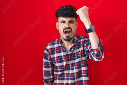 Young hispanic man with beard standing over red background angry and mad raising fist frustrated and furious while shouting with anger. rage and aggressive concept.
