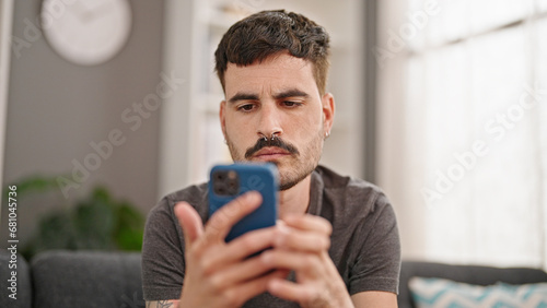 Young hispanic man using smartphone sitting on sofa at home