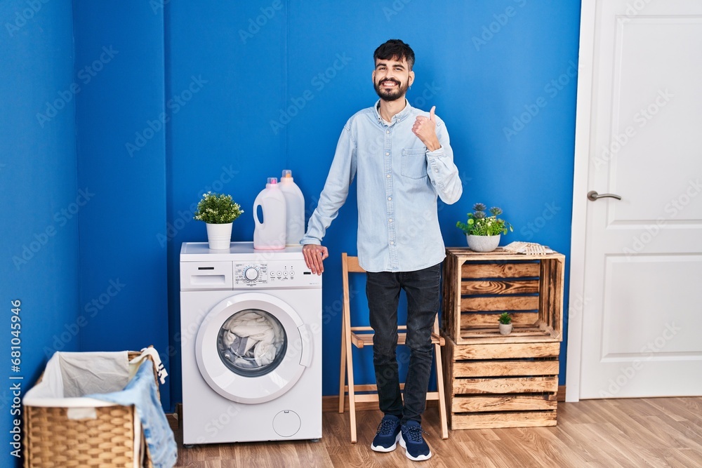 Young hispanic man with beard doing laundry standing at laundry room smiling happy and positive, thumb up doing excellent and approval sign Young hispanic man with beard doing laundry standing at laundry room smiling happy and positive, thumb up doing excellent and approval sign