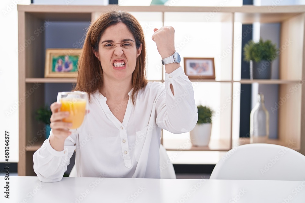Brunette woman drinking glass of orange juice angry and mad raising fist frustrated and furious while shouting with anger. rage and aggressive concept. Brunette woman drinking glass of orange juice angry and mad raising fist frustrated and furious while shouting with anger. rage and aggressive concept.