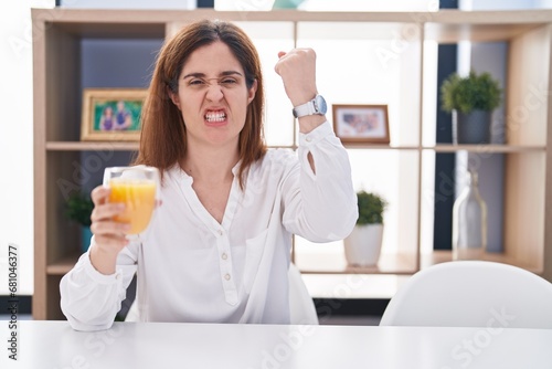 Brunette woman drinking glass of orange juice angry and mad raising fist frustrated and furious while shouting with anger. rage and aggressive concept.