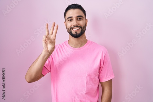 Hispanic young man standing over pink background showing and pointing up with fingers number three while smiling confident and happy.