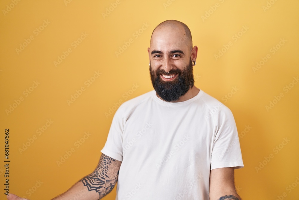 Young hispanic man with beard and tattoos standing over yellow background smiling cheerful with open arms as friendly welcome, positive and confident greetings Young hispanic man with beard and tattoos standing over yellow background smiling cheerful with open arms as friendly welcome, positive and confident greetings