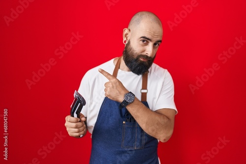 Young hispanic man with beard and tattoos wearing barber apron holding razor pointing with hand finger to the side showing advertisement, serious and calm face © Krakenimages.com